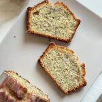 Two slices and a loaf of iced lemon poppy seed bread on a white plate with a fresh lemon.
