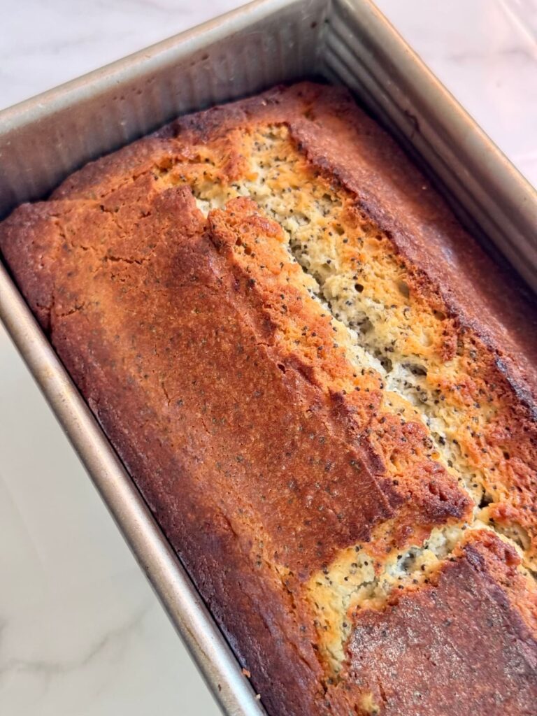 A close-up, angled view of a freshly baked lemon poppy seed loaf with a golden-brown crust, still in a rectangular metal baking pan.