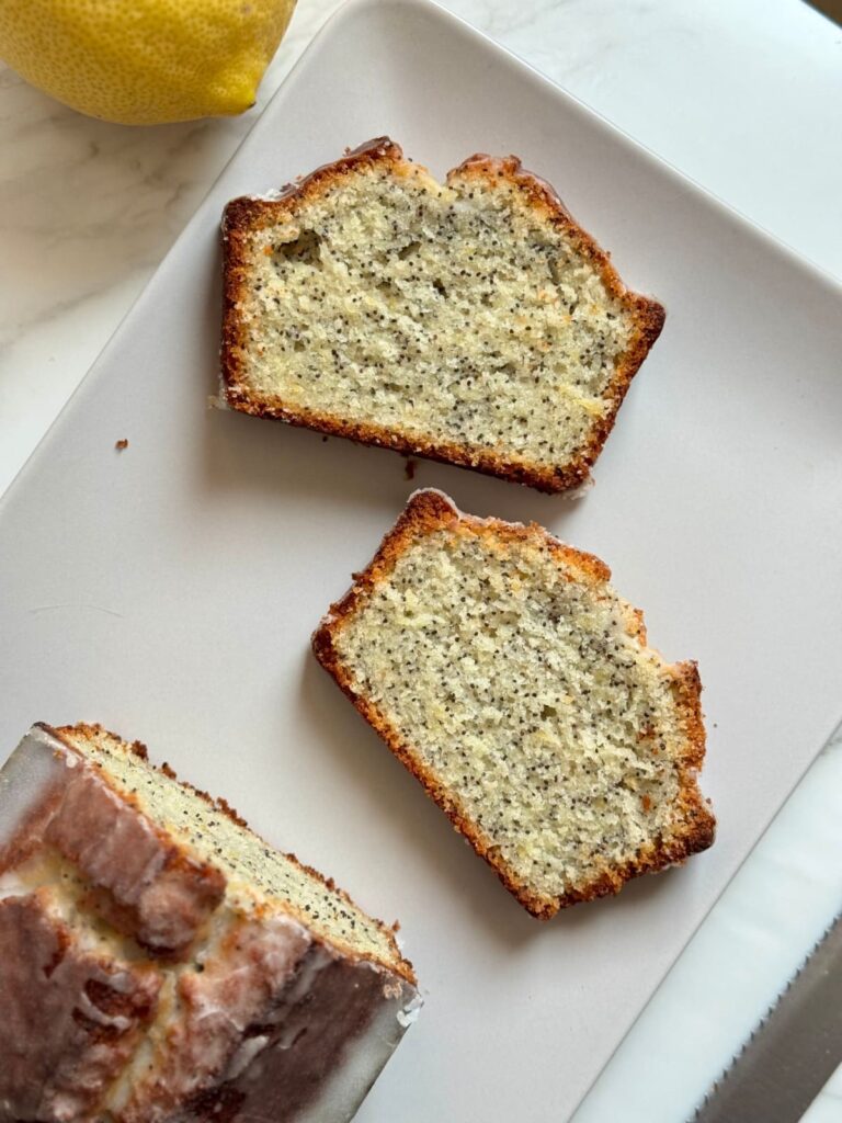 Two slices and a loaf of iced lemon poppy seed bread on a white plate with a fresh lemon.