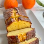 A sliced loaf of iced orange bread on a white rectangular platter, with two whole oranges in the background.