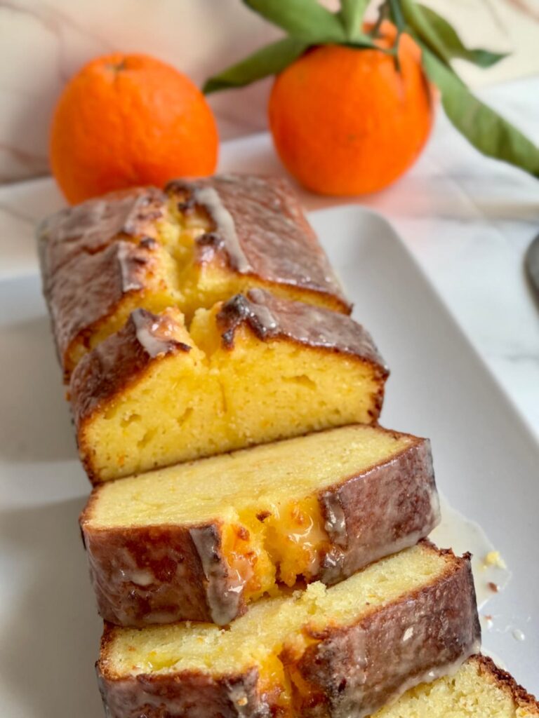 A sliced loaf of iced orange bread on a white rectangular platter, with two whole oranges in the background.