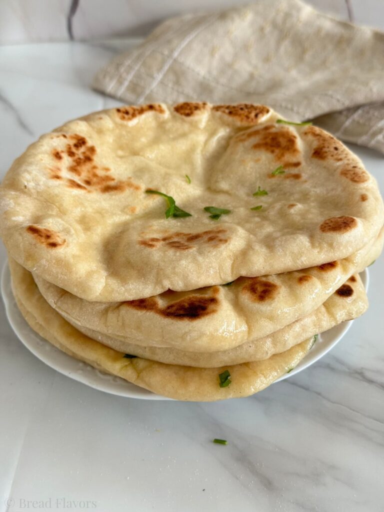 A close-up stack of three warm, pillowy naan breads on a white plate, showing golden-brown charred spots, brushed with butter and topped with a parsley garnish.