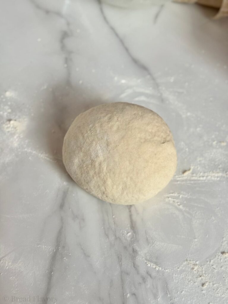 A smooth, round ball of 2 ingredient naan dough resting on a lightly floured marble countertop.