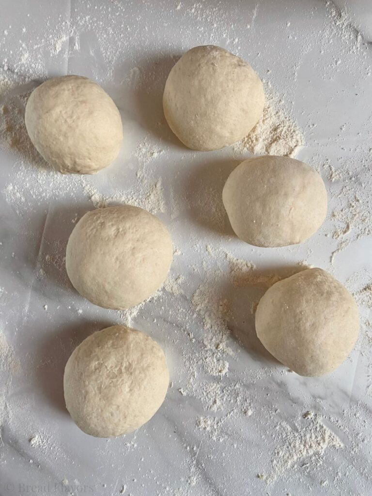Six small, round portions of naan dough arranged on a floured marble surface, ready to be rolled out.