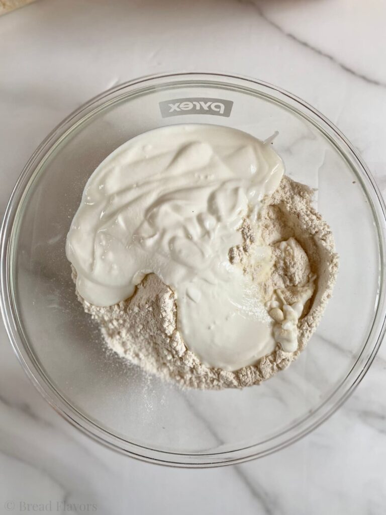 A glass mixing bowl containing white flour topped with a thick layer of Plain yogurt.