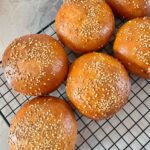 Overhead view of six golden-brown, round semolina buns topped with sesame seeds, cooling on a black wire rack.