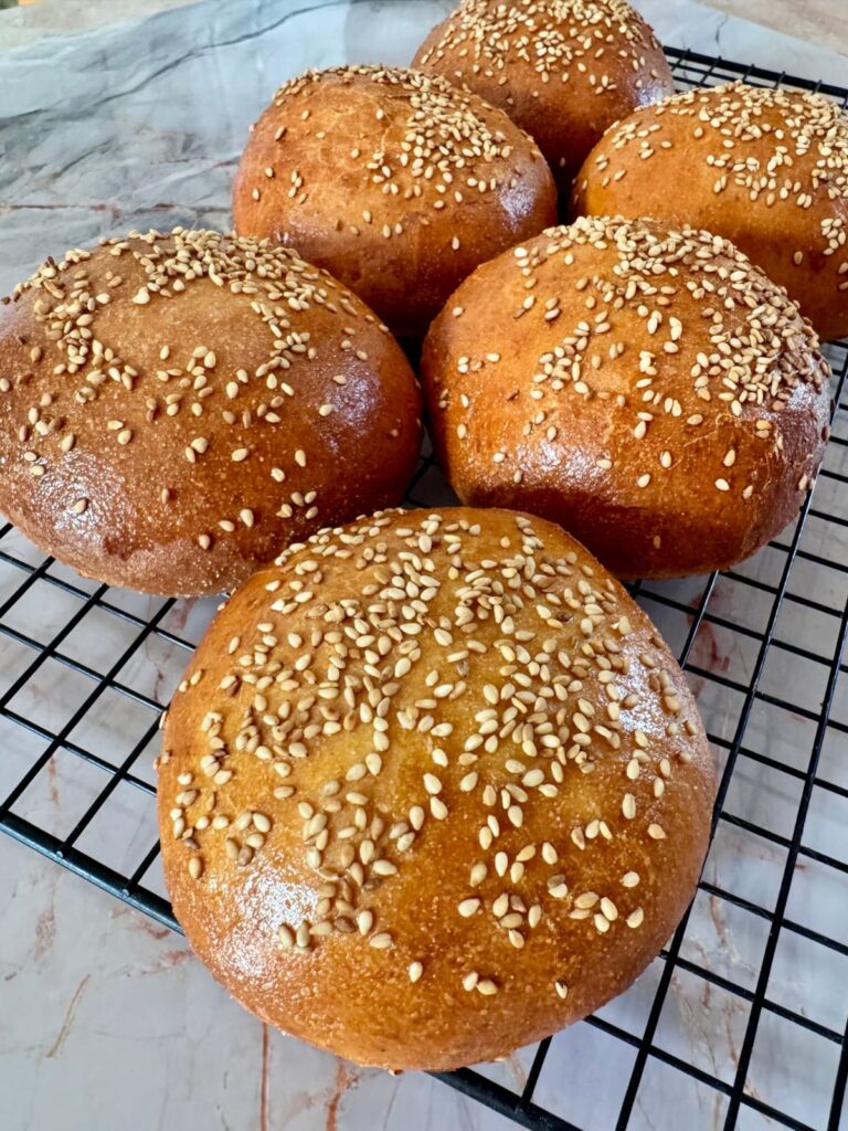 Close-up, angled shot of several shiny, sesame-crusted semolina buns resting on a black cooling rack.
