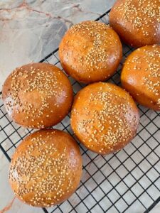 Overhead view of six golden-brown, round semolina buns topped with sesame seeds, cooling on a black wire rack.