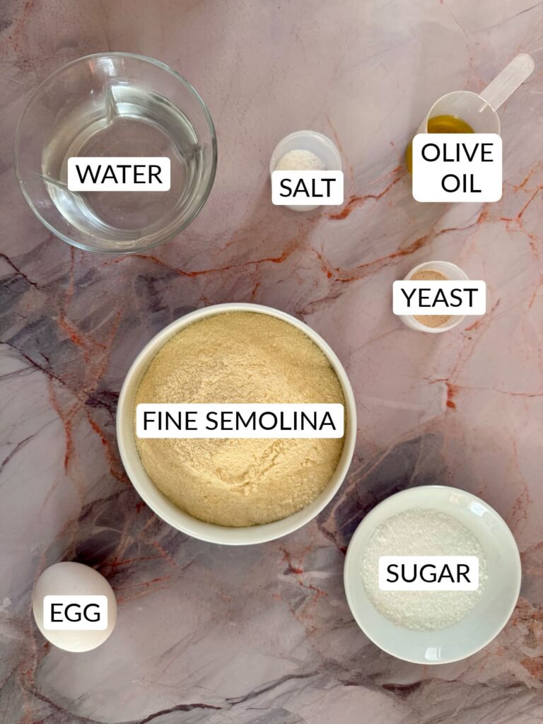 Top-down view of labeled ingredients: fine semolina, water, salt, olive oil, yeast, sugar, and an egg.