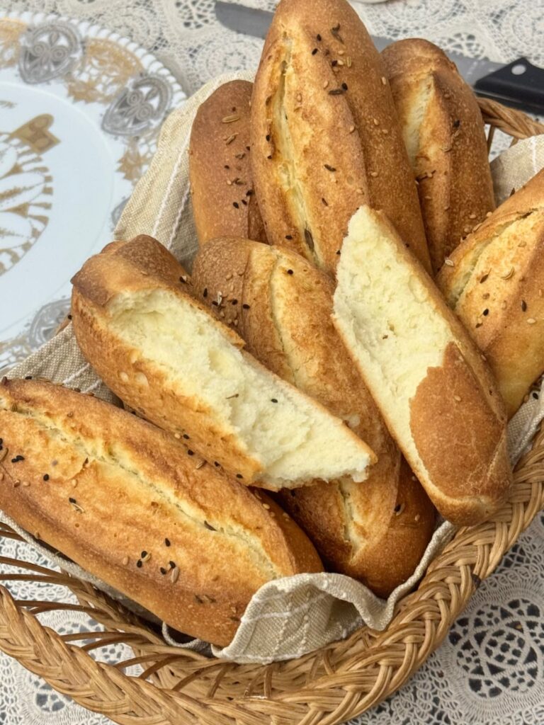 A wicker basket filled with golden-brown semolina rolls topped with sesame seeds, black sesame seeds, and fennel seeds.