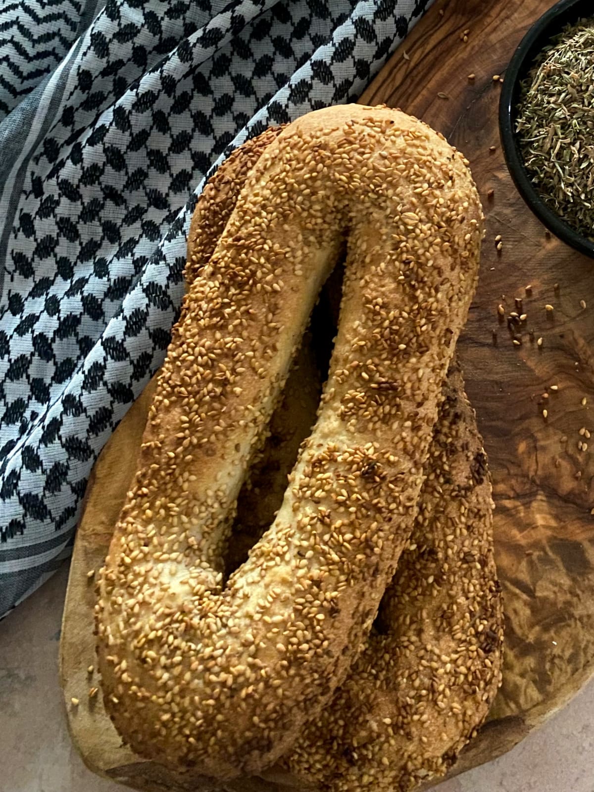 wo sesame-crusted Jerusalem bagels stacked on a wooden board next to a keffiyeh.