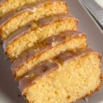 A side-angle close-up of a moist lemon breadloaf, sliced into thick pieces and arranged on a light grey platter.