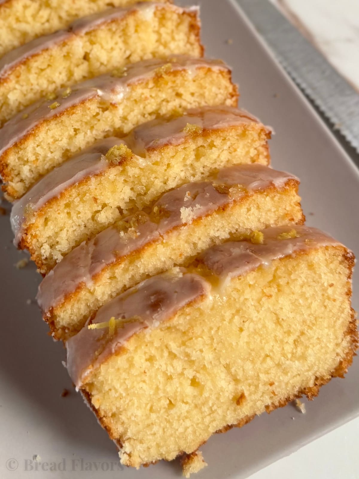A side-angle close-up of a moist lemon breadloaf, sliced into thick pieces and arranged on a light grey platter.