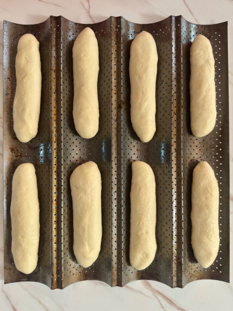 Eight shaped dough logs resting on a perforated baguette pan before their final rise.