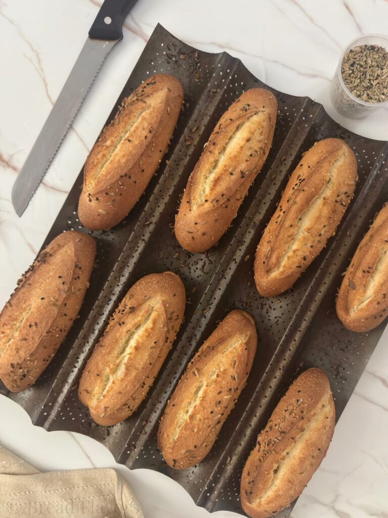 Eight fresh baked semolina rolls cooling on a baguette pan next to a bread knife.